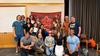 Indoor group photo of 17 smiling adults in front of red tapestry, some holding plush toys