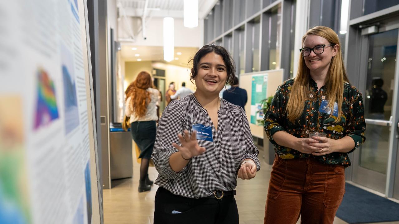 Two smiling women in a bright gallery hallway, one holding a blue paper hand.