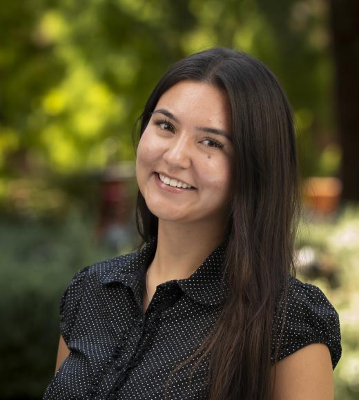 Young woman with long dark hair smiling in outdoor headshot, blurred green background