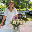 Smiling woman in white blouse behind table with glass vase of white and pink flowers, outdoor photo